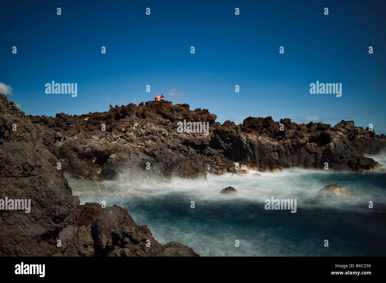 Shoreline, Los Cancajos. La Palma. Die Lava Rock Küsten Relief, die typisch für die Insel sind. Zwei Touristen sitzen auf einer öffentlichen Bank hoch oben auf den Felsen, die Aussicht zu genießen. Es ist ein Sonniger Tag mit einem blauen Himmel. Stockfoto