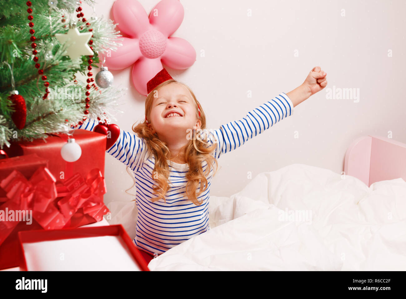 Glückliches Kind Mädchen in santa hut zieht die Hände oben sitzen im Bett nach dem Aufwachen. Weihnachten Morgen. Stockfoto