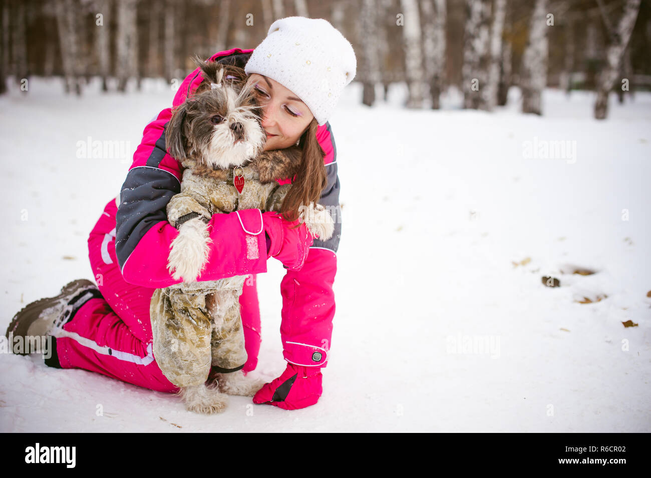 Spaziergang im Winter im Freien mit Hund Rasse Shih Tzu. Eine Frau in leuchtend roten warme Skibekleidung Wandern im Schnee mit Ihrem Haustier, kleine Shih Tzu in Overalls gekleidet. Pflege für Tiere liebt das Spiel mit dem Hund Stockfoto