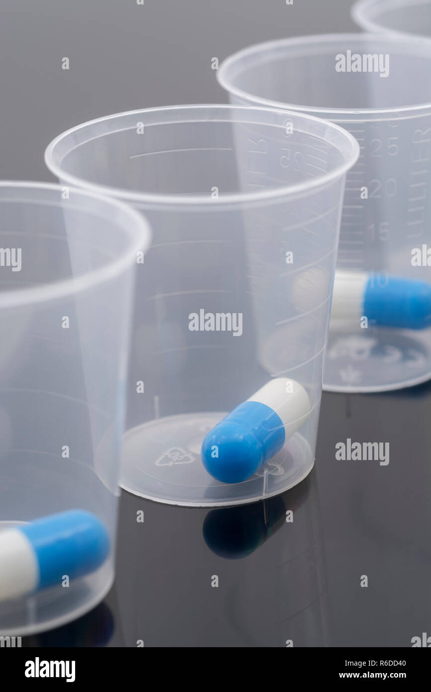 Big Pharma Medikamente Konzept. Blaue und Weiße Kapseln Pillen in Kunststoff Messbecher / messbecher auf reflektierende Oberfläche. Möglich NHS Metapher. Stockfoto