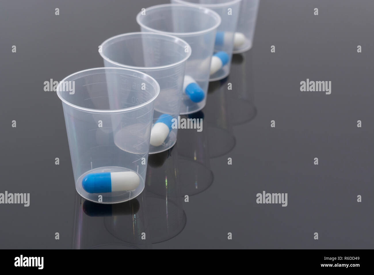 Big Pharma Medikamente Konzept. Blaue und Weiße Kapseln Pillen in Kunststoff Messbecher / messbecher auf reflektierende Oberfläche. Möglich NHS Metapher. Stockfoto