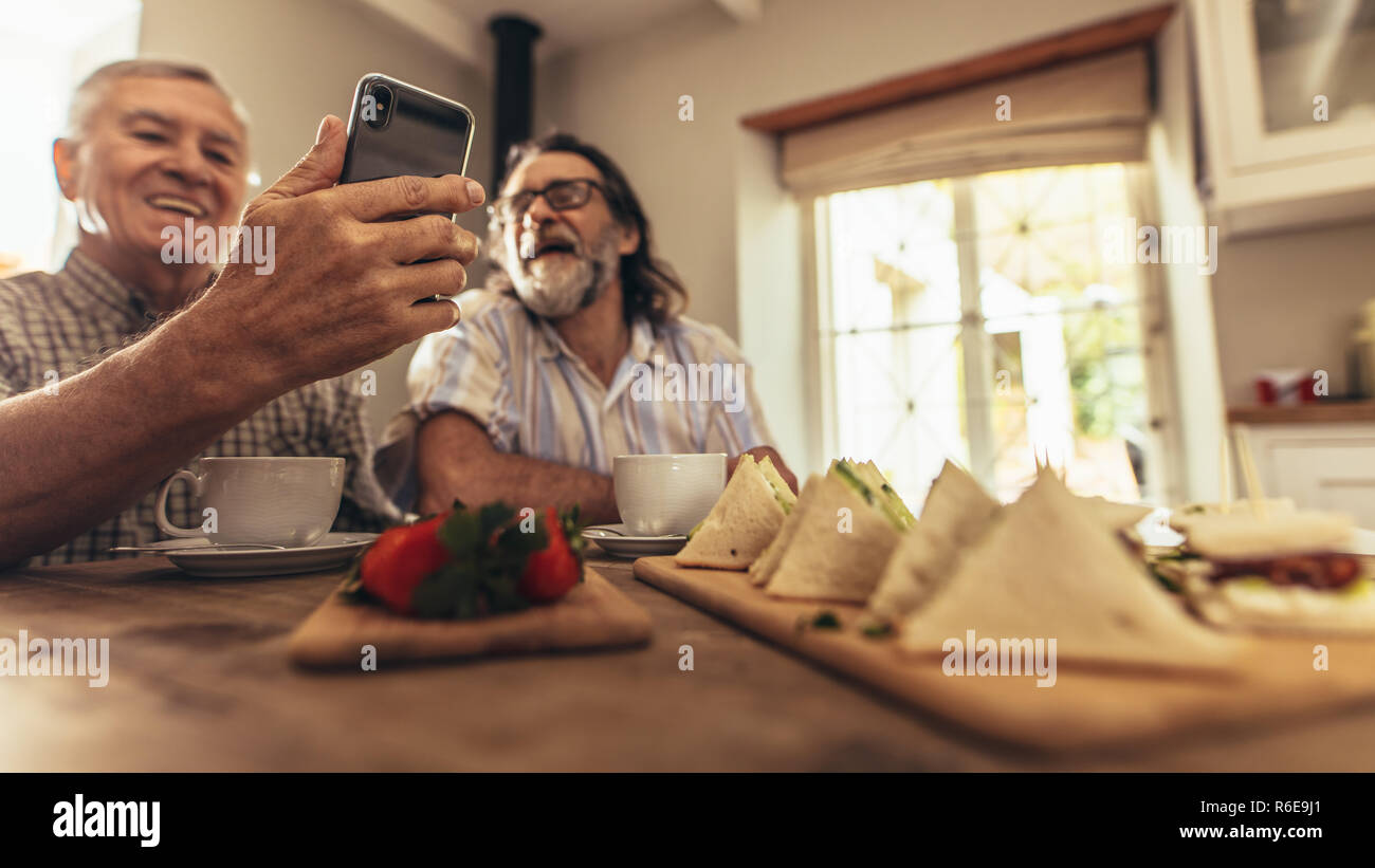 Ältere Männer, die ein Video mit Freunden auf dem Smartphone chatten. Fokus auf ältere männliche Hände halten Handy am Tisch drinnen sitzen. Stockfoto