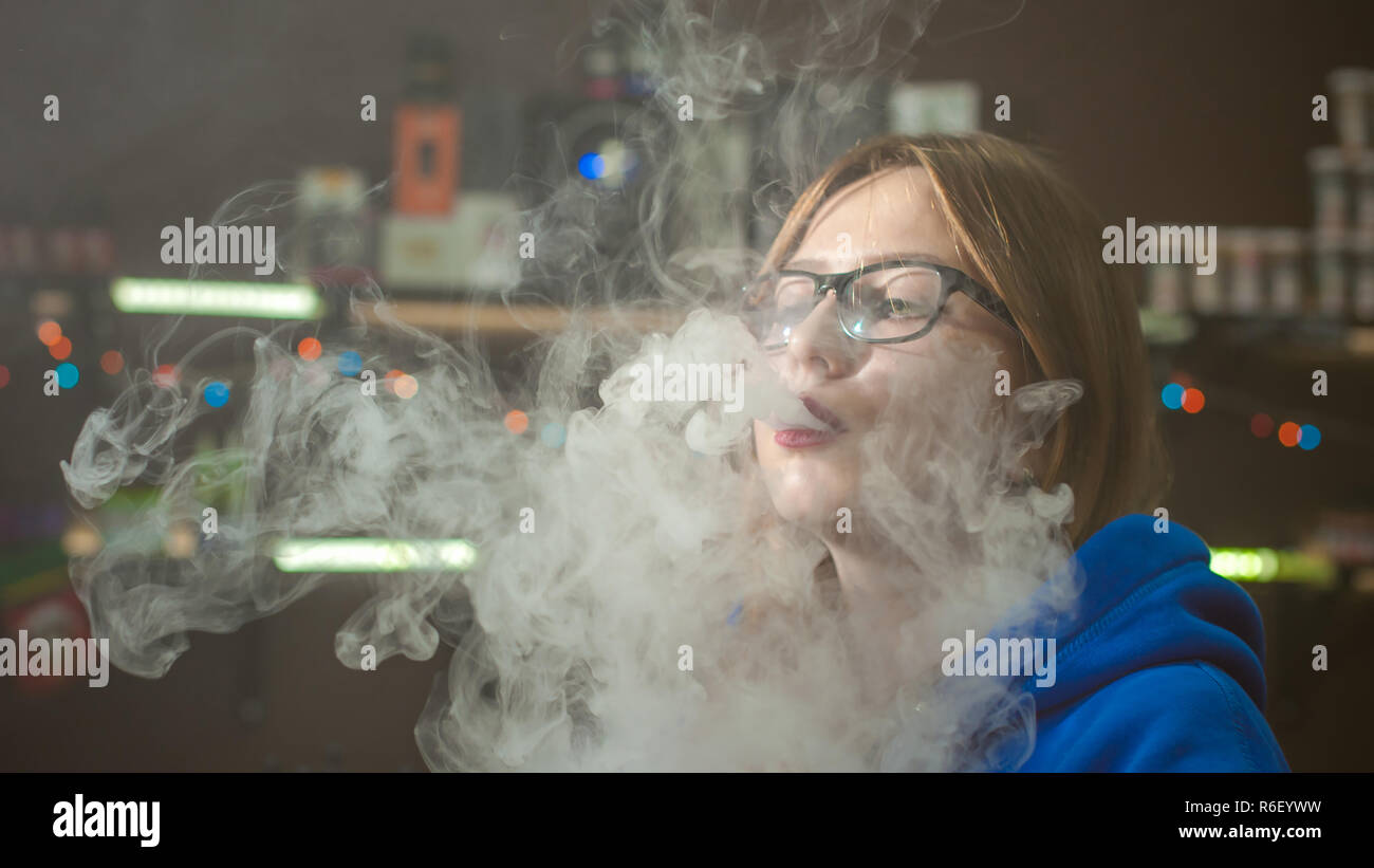 Schöne junge Frau mit Brille rauchen Elektronische Zigarette. Mädchen in  einem blauen Pullover weht Wolken von Glycerin Dampf, von vape Mod, sitzen drinnen  vape Shop Cafe Stockfotografie