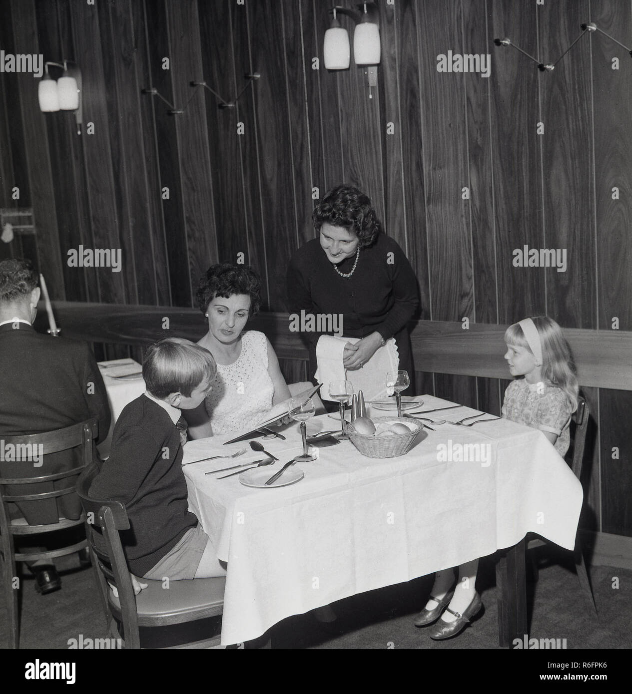 1960er Jahre, historisch, zwei kleine Kinder sitzen mit ihrer Mutter an einem Tisch in einem holzgetäfelten Feinschmeckerrestaurant und schauen sich die Speisekarte an, während sie von einer Kellnerin aus England, Großbritannien, serviert werden. Stockfoto