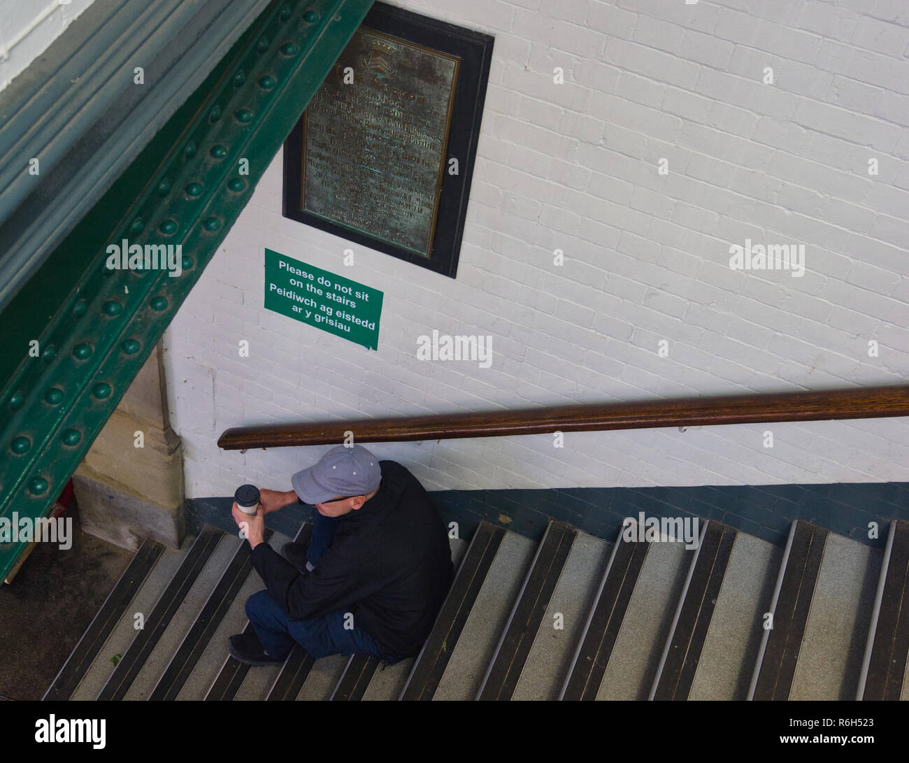 Mann sitzt auf der nächsten Schritte in Englisch und Walisisch zu Schild mit der Aufschrift "Bitte nicht auf der Treppe", Cardiff, Cardiff, Wales, Vereinigtes Königreich sitzen Stockfoto