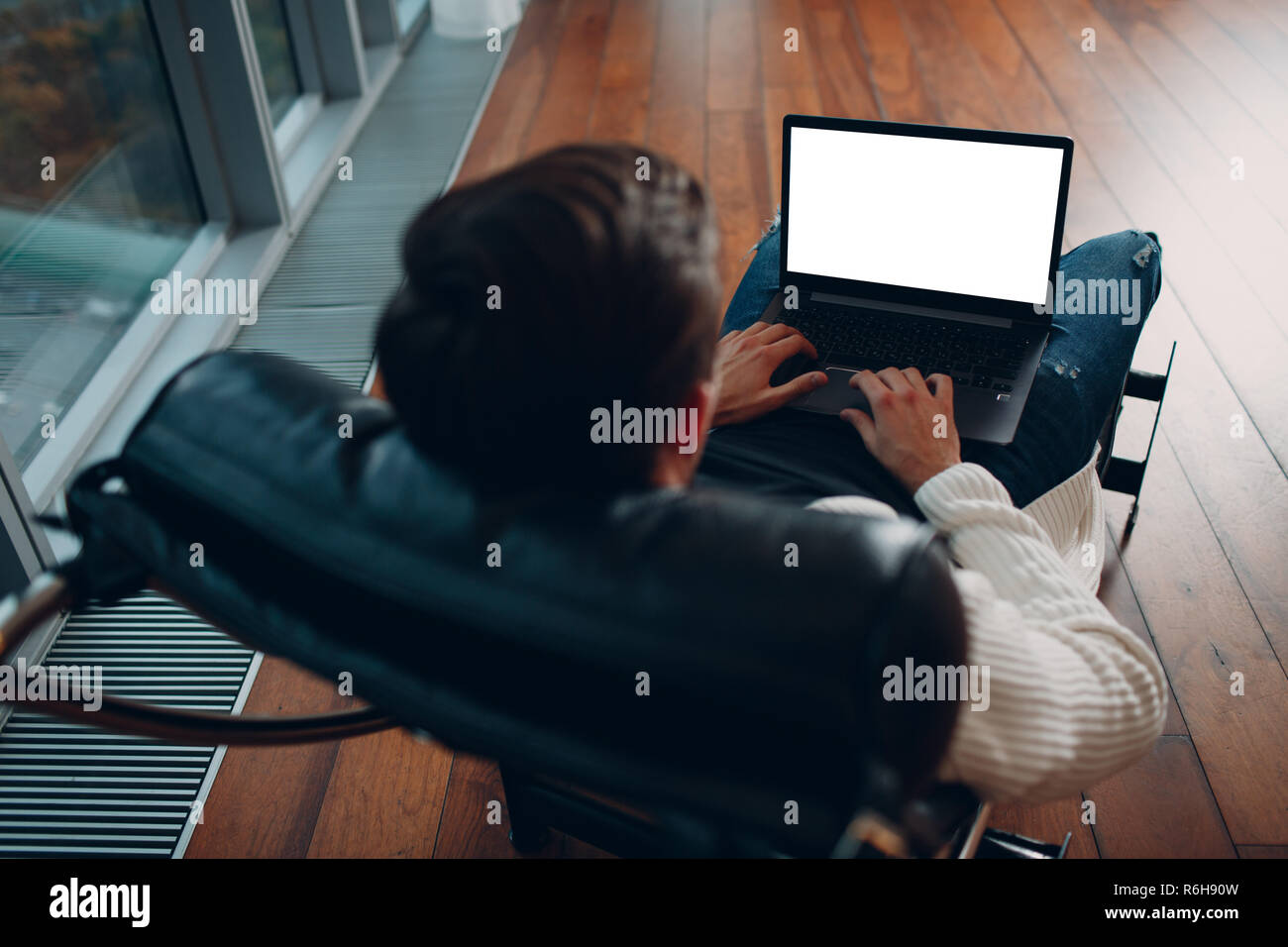 Ein junger Mann mit Laptop mit leeren weißen Bildschirm sitzt auf einem Stuhl am Fenster in einem Wolkenkratzer. Stockfoto
