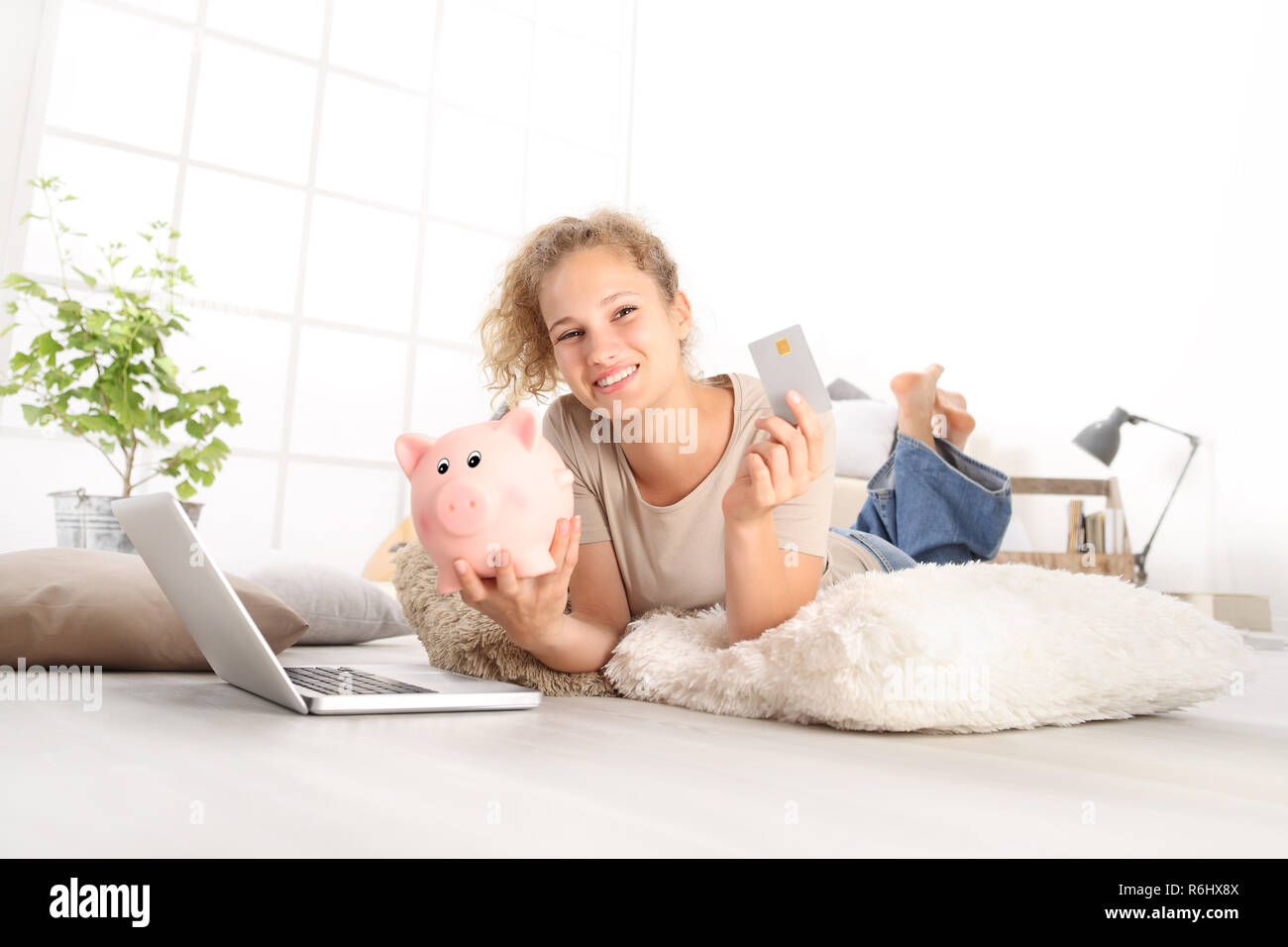 Junge lächelnde Frau mit Kreditkarte und Sparschwein, mit Laptop Computer online shopping sparen Konzept, liegen auf Wohnzimmer Holzboden in Co Stockfoto