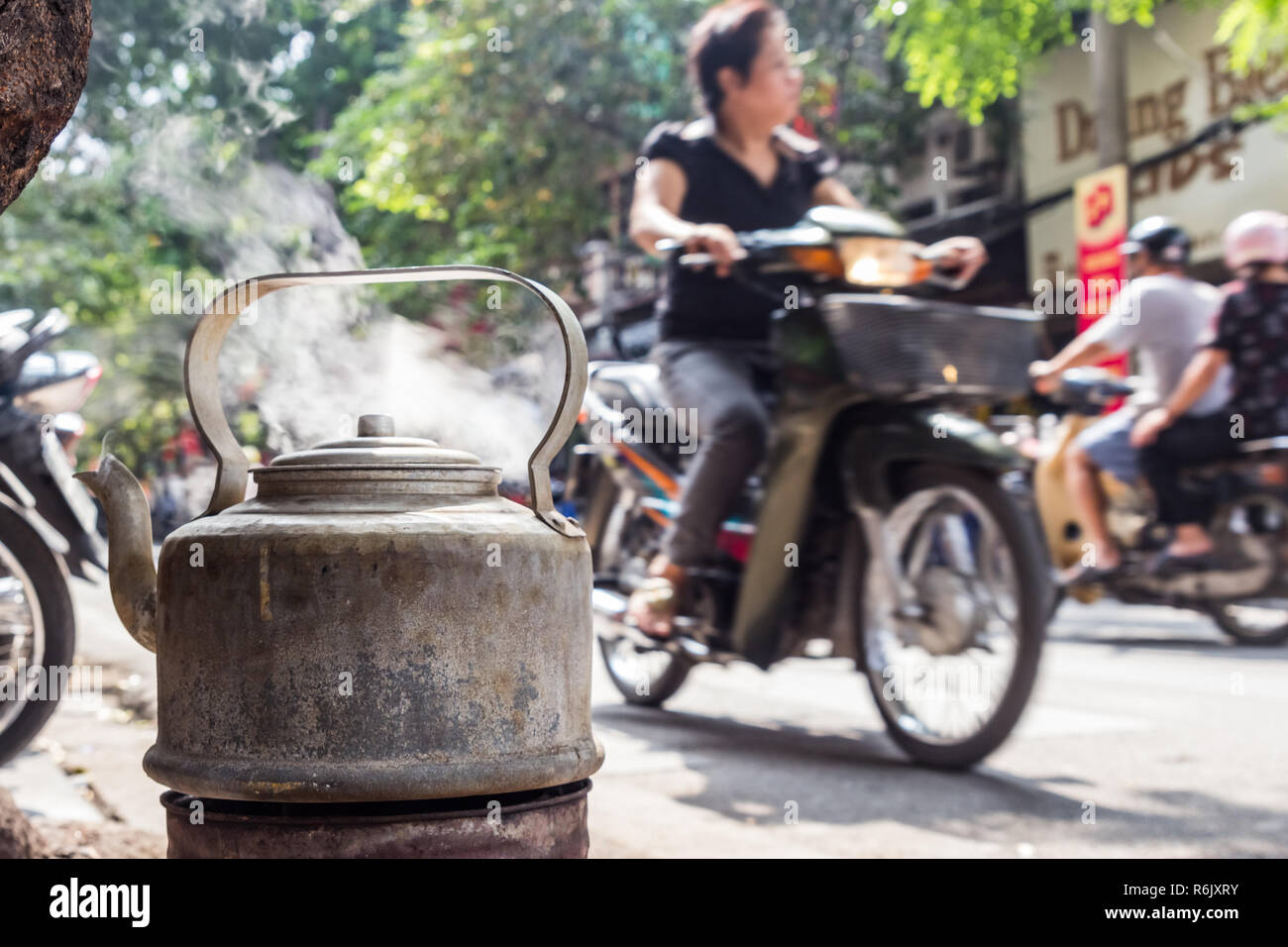 Kochendes Wasser in alten Wasserkocher aus Metall auf der Straße in Hanoi, Vietnam. Hintergrund verschwommen, nicht erkennbare Frau auf dem Motorroller. Stockfoto