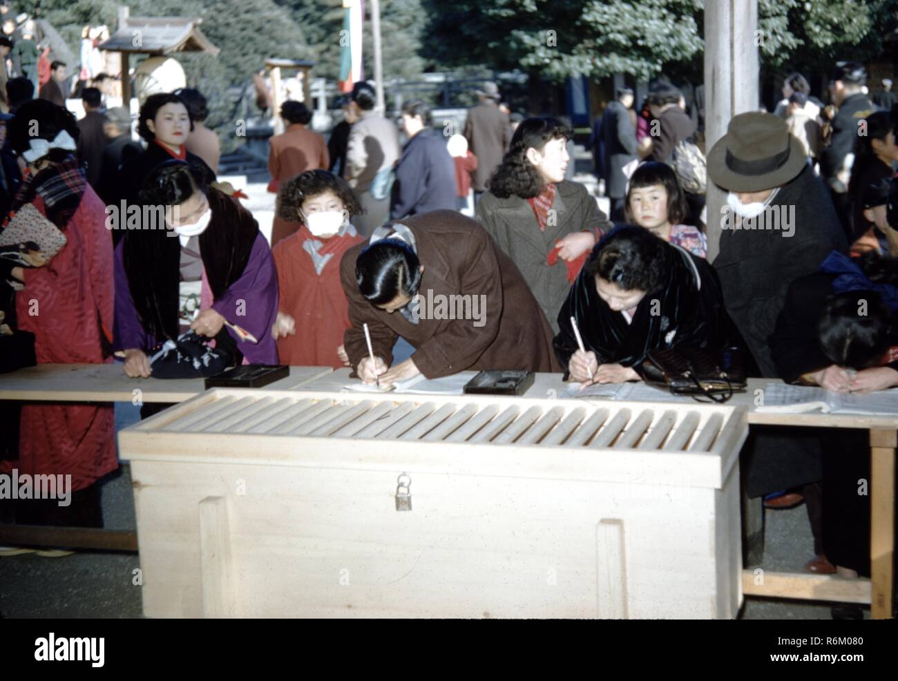 Farbfoto, die japanische Männer, Frauen und Kinder, in einer Parkähnlichen, einige tragende traditionellen Kimonos und andere Mäntel tragen, mit ein paar Leute im Vordergrund schiefen über einen Zähler und Schreiben in der geöffneten Seiten des Booklets und eine gesperrte White Box oder Kühler im Vordergrund, wahrscheinlich in Japan während der Mitte des zwanzigsten Jahrhunderts, 1965 fotografiert. () Stockfoto