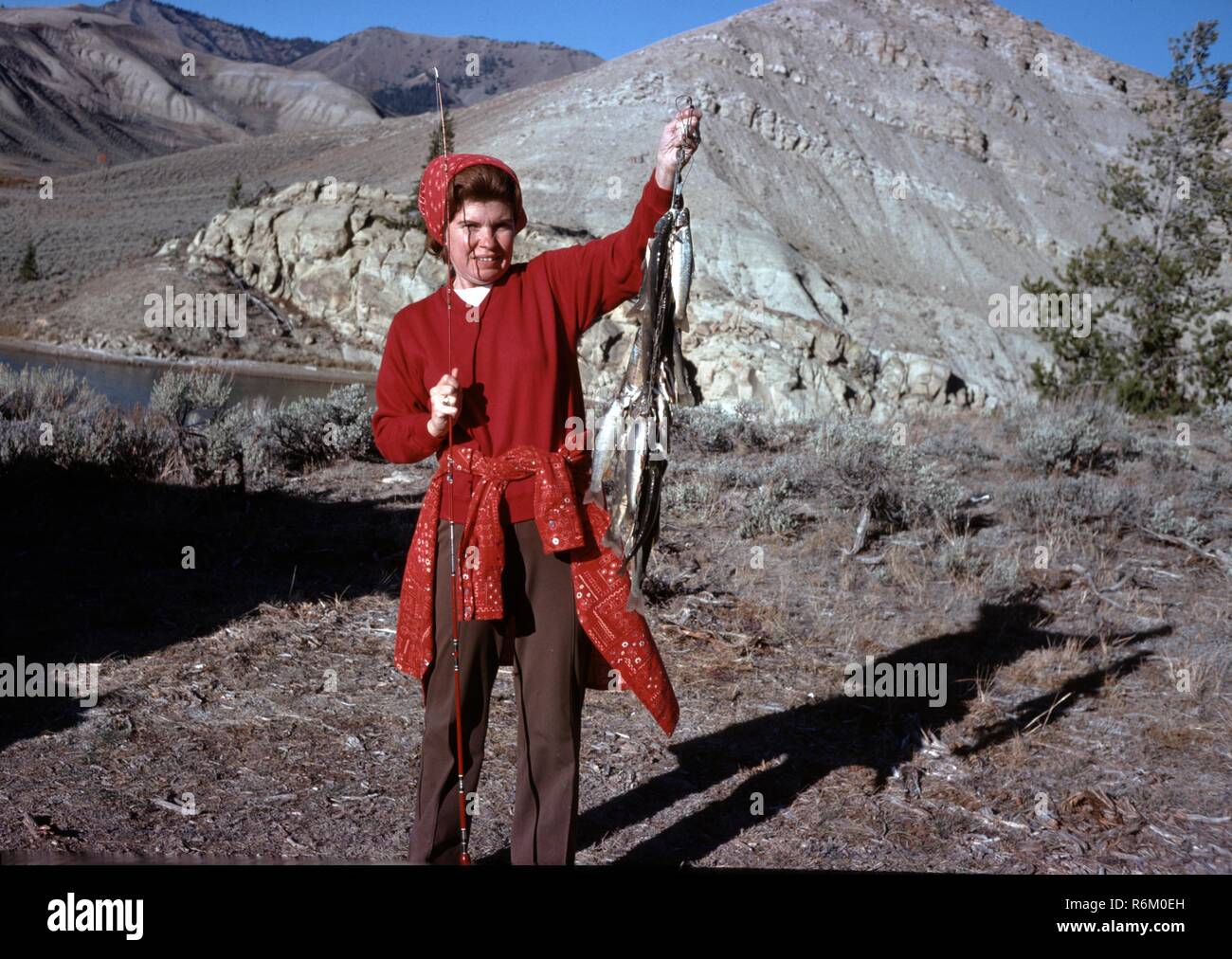 Junge Frau in rotem Hemd und Schal stolz Fang kleiner Fische gekleidet, mit Angelrute in Ihrer anderen Hand, mit Blick auf den See und die Berge im Hintergrund, Wyoming, 1976. () Stockfoto