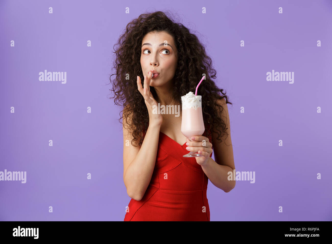 Porträt einer flirty Frau mit dunklen lockigen Haar rotes Kleid über violett Hintergrund isoliert, trinken Milch aus einem Glas schütteln Stockfoto