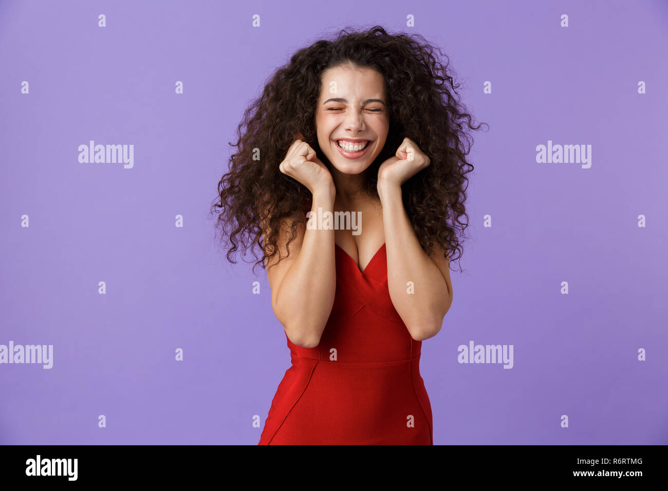 Porträt eines fröhliche Frau mit dunklen lockigen Haar rotes Kleid über violett Hintergrund isoliert, Erfolg feiern. Stockfoto