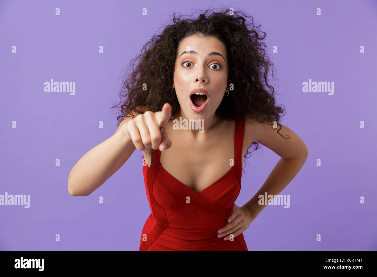 Portrait von eine aufgeregte Frau mit dunklen lockigen Haar rotes Kleid über violett Hintergrund isoliert, Zeigefinger an Kamera Stockfoto
