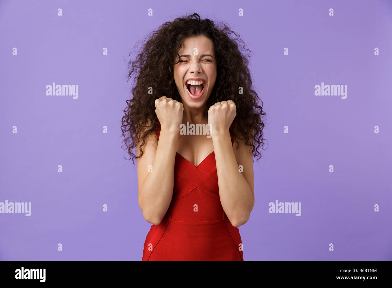 Porträt eines fröhliche Frau mit dunklen lockigen Haar rotes Kleid über violett Hintergrund isoliert, Erfolg feiern. Stockfoto