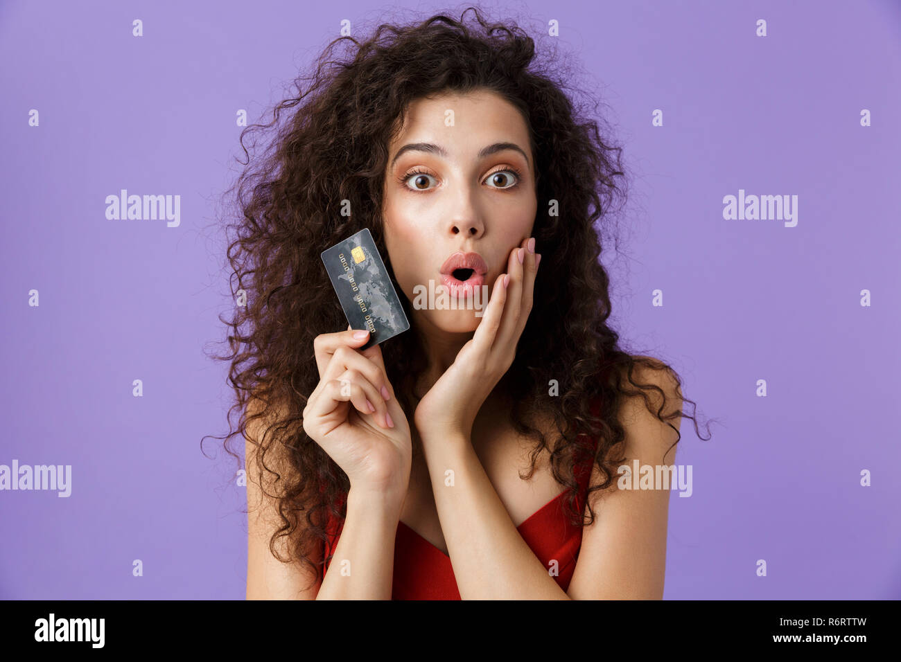 Portrait von eine aufgeregte Frau mit dunklen lockigen Haar rotes Kleid über violett Hintergrund isoliert, Kunststoff Kreditkarte Stockfoto