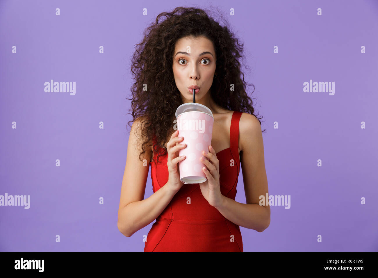 Portrait einer hübschen frau mit dunklen lockigen Haar rotes Kleid über violett Hintergrund isoliert, trinken aus einem Plastikbecher mit Strohhalm Stockfoto