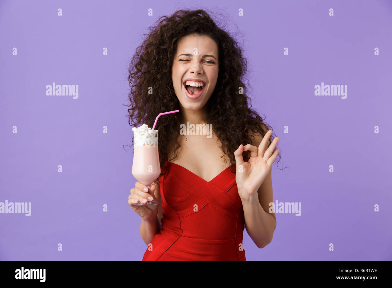 Portrait von freut sich Frau mit dunklen lockigen Haar rotes Kleid über violett Hintergrund isoliert, trinken Milch shake aus einem Glas, ok angezeigt Stockfoto