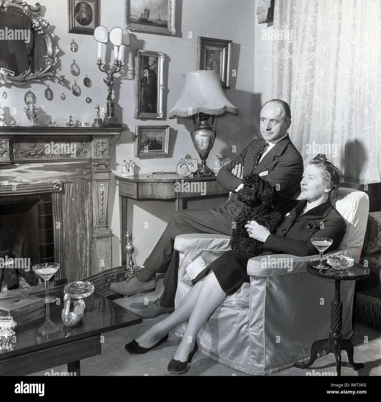 1950, historische, eine englische Dame in einem Sessel in der Lounge bei einem Drink sitzen und Ihr Haustier Hund Holding, mit ihrem Mann neben ihr sitzen auf dem Stuhl Arme, England, UK. Stockfoto