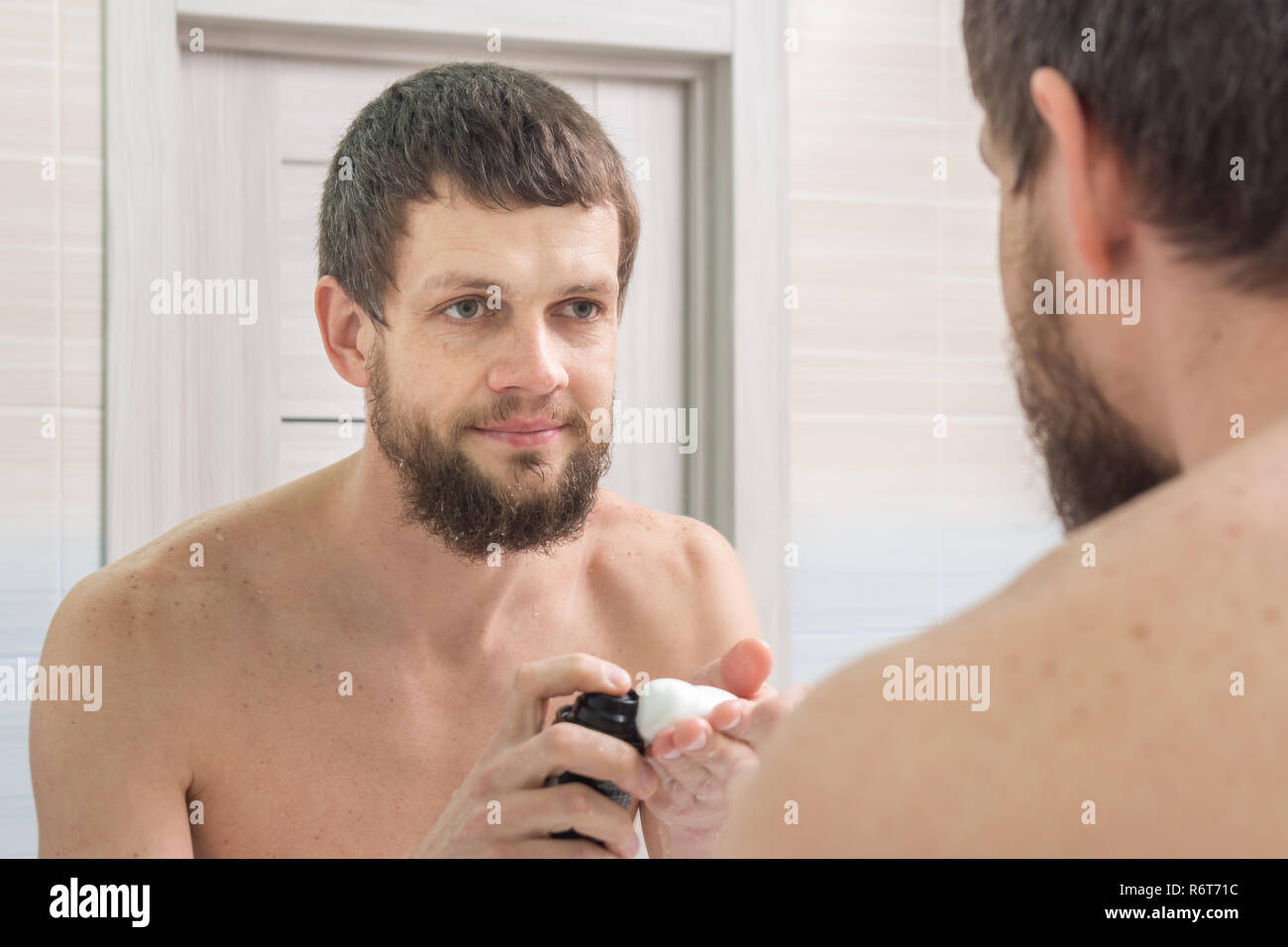 Einen unrasierten Mann gilt einen Rasierschaum auf die Hand und schaut in den Spiegel im Badezimmer Stockfoto