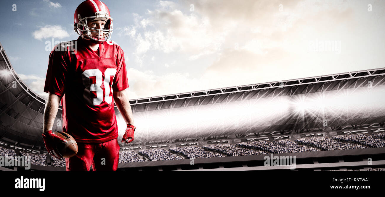 American football player stehend mit rugby ball und Helm gegen überfüllten Stadion mit bewölktem Himmel Stockfoto