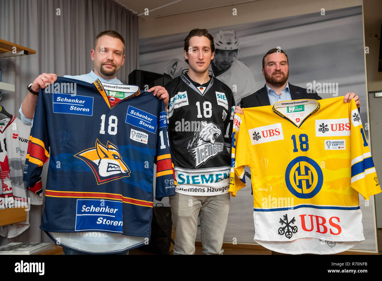 Nürnberg, Bayern, Deutschland. 7. Dezember 2018. Anton Chiaka (L-R), Stellvertretender Geschäftsführer der Russischen Eishockey Club Magnitogorsk, Brandon Buck, Stürmer der Thomas Sabo Ice Tigers und Marc Gianola, Präsident des Sprengler Schale, stehen Seite an Seite mit Trikots der teilnehmenden Mannschaften am Spengler Cup 2018 während der Pressekonferenz für das Turnier. Der Spengler Cup 2018 findet vom 26.-31.12.2018 in Davos (Schweiz). Quelle: dpa Picture alliance/Alamy leben Nachrichten Stockfoto