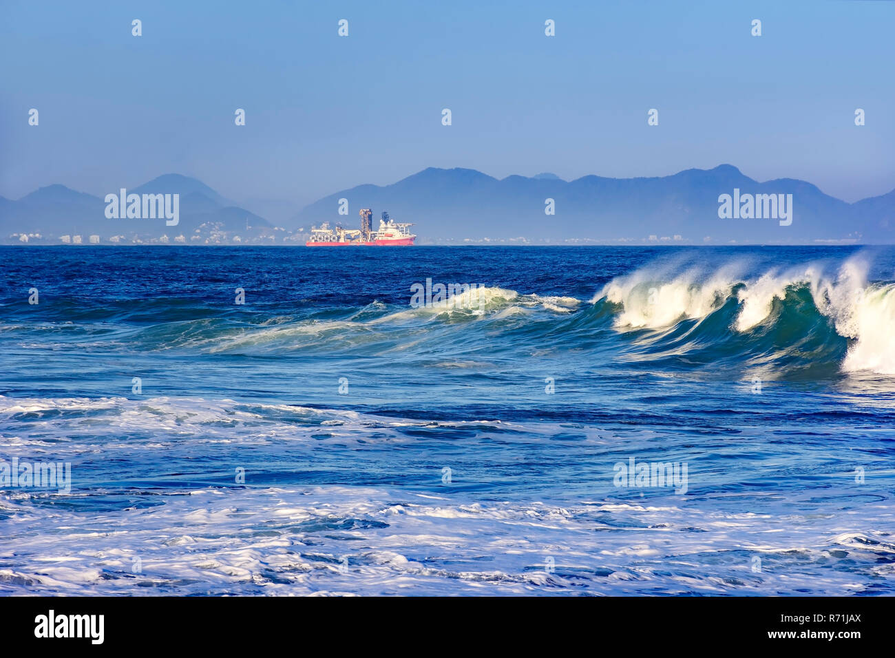 Wellen brechen am Strand des Teufels in Rio de Janeiro mit Frachtschiff, die Stadt und die Hügel im Hintergrund Stockfoto