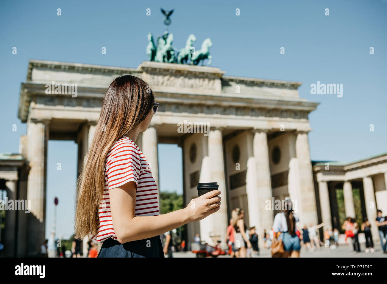 Eine Person hält eine Tasse mit Kaffee oder einem anderen Getränk beim Gehen oder Sightseeing in Berlin, Deutschland. Die Zukunft ist das Brandenburger Tor und verschwommenes unbekannte Menschen. Stockfoto