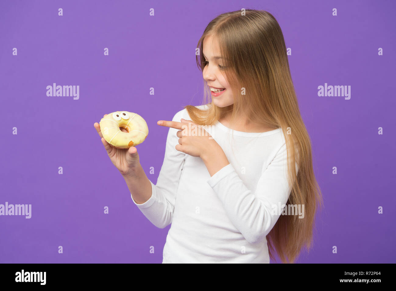 Glückliches Kind zeigen mit dem Finger auf Krapfen auf Violett Hintergrund. Kleines Mädchen mit glasierten ring Donut auf lila Hintergrund. Das ungesunde Essen. Kind Lächeln mit Junk Food. Essen und Nachtisch. Kindheit und Kinderbetreuung. Stockfoto