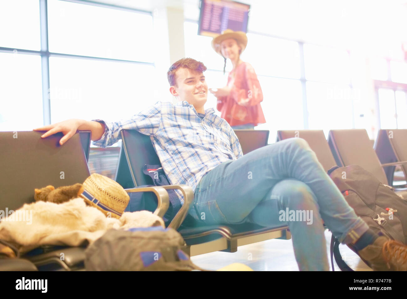 Junger Mann am Flughafen sitzen, Rucksack neben sich, junge Frau, die in der Nähe stehen, auf ihn zu schauen, Stockfoto