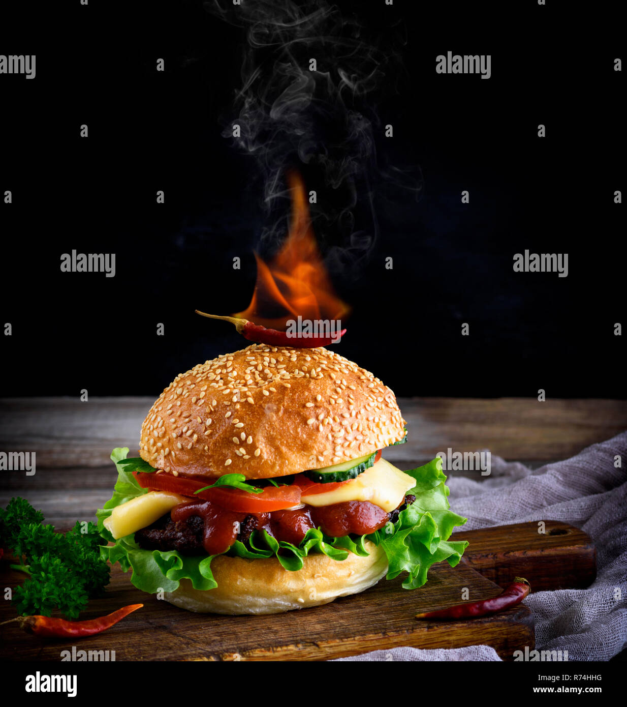 Classic Burger mit einer Frikadelle, Käse und Gemüse, oben auf einem Brötchen mit einem Sesam ist eine brennende rote Chilischote Stockfoto