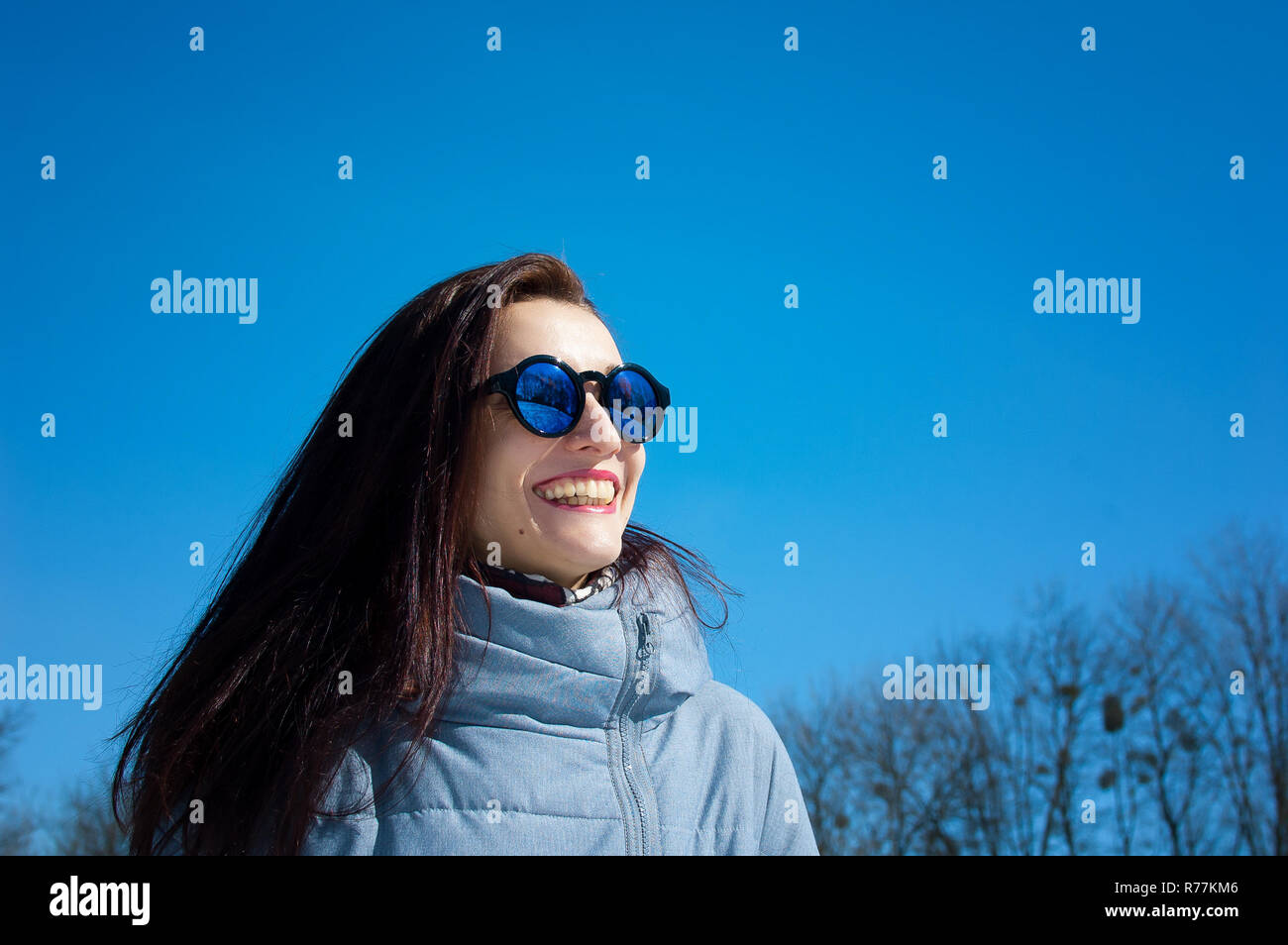 Im freien Lebensstil Nahaufnahme Portrait von schönen Mädchen zu Fuß in den schneereichen Winter Park. Lächelnd und Winter genießen. Das Tragen von eleganten verspiegelten Sonnenbrillen, blau Trenchcoat Stockfoto