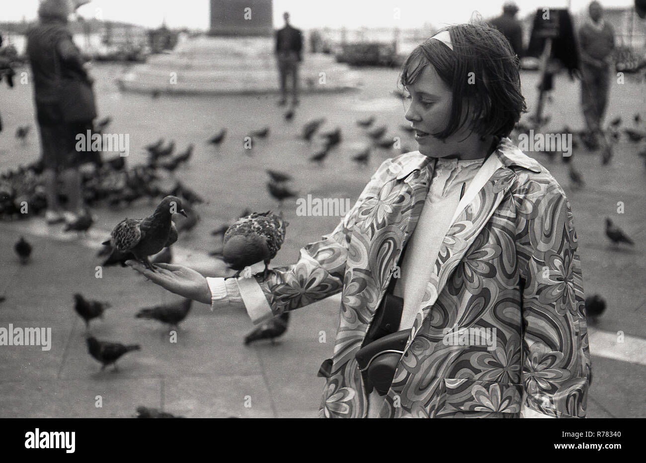 Siebziger Jahre, zwei Tauben auf dem Arm eines jungen Mädchens in einem blumengemusterten Mantel, der auf dem Markusplatz (Markusplatz) in Venedig, Italien, steht. Tauben waren schon immer eine beliebte Touristenattraktion in Venedig und konkurrierten einst mit Katzen als die traditionellen, wenn auch inoffiziellen Maskottchen der Lagunenstadt. Stockfoto