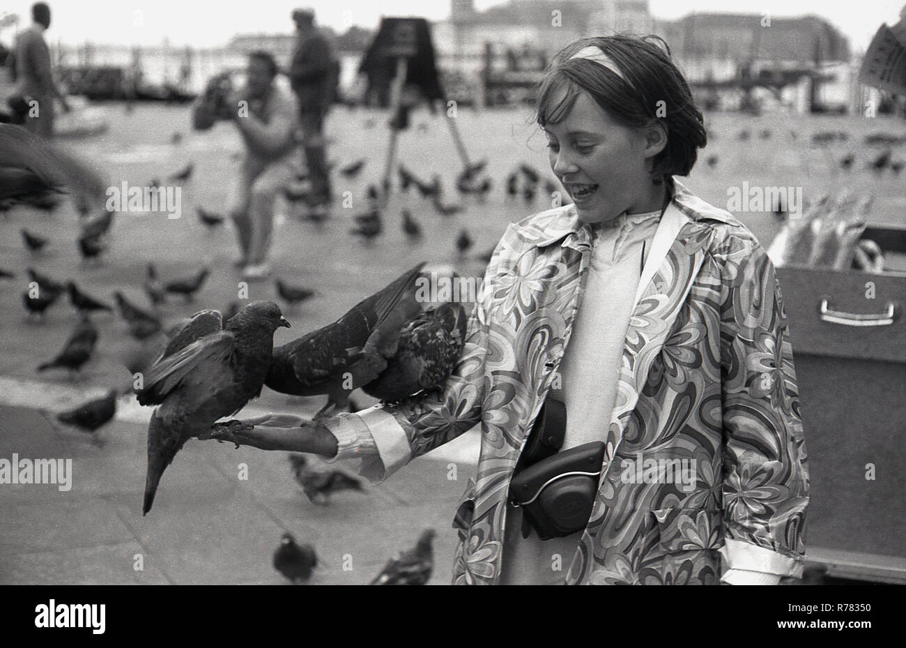 Siebziger Jahre, Tauben auf dem Arm eines jungen Mädchens in einem gemusterten Mantel, das auf dem Markusplatz oder dem Markusplatz in Venedig, Italien, steht. Tauben waren schon immer eine beliebte Touristenattraktion in Venedig und konkurrierten einst mit Katzen als die traditionellen, wenn auch inoffiziellen Maskottchen der Lagunenstadt. Stockfoto