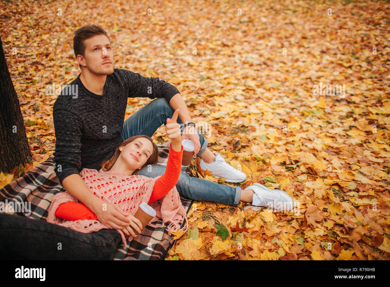 Schönes Paar sitzen und im Herbst Park allein liegen. Sie schauen in die gleiche Richtung. Liegende Frau auf die Beine Kerl. Kerl sitzt auf Decke. Sie halten Tasse Kaffee. Stockfoto