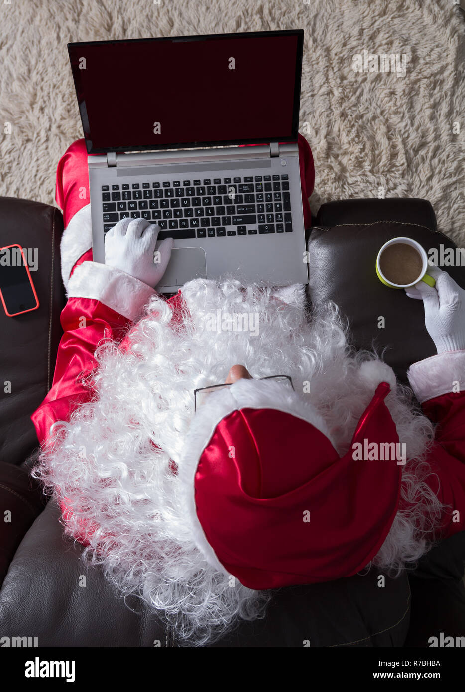 Overhead von Santa Claus (oder Kris Kringle) Entspannung im Sofa zu Hause. Mit Laptop für Kommunikation und Freizeit- und trinken Kaffee mit Milch. Stockfoto