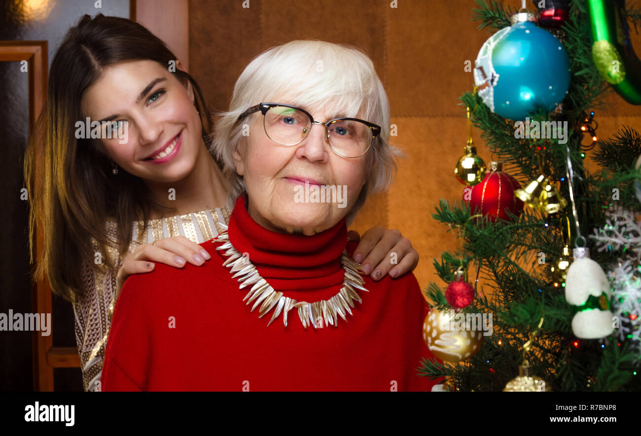 Für das neue Jahr. senior Oma und jungen kaukasischen Frau, Teenager in der Nähe von Weihnachten Baum. Enkelin, jugendlich umarmt, umarmt alte Großmutter. festliche Stimmung Stockfoto