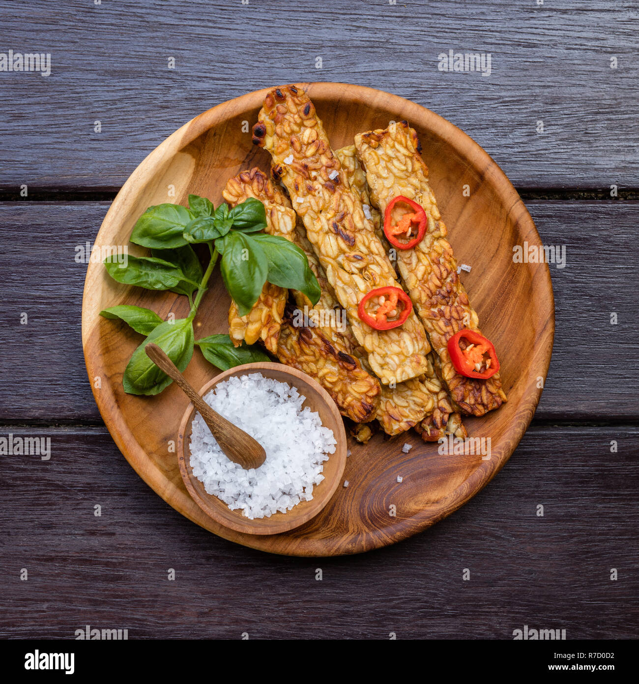 Gebratenen tempeh mit Chili Pfeffer und Basilikum, Meersalz in Holz- Topf mit Löffel aus Holz auf einem runden Holz- fach über der Oberfläche aus dunklem Holz eingerichtet. Stockfoto