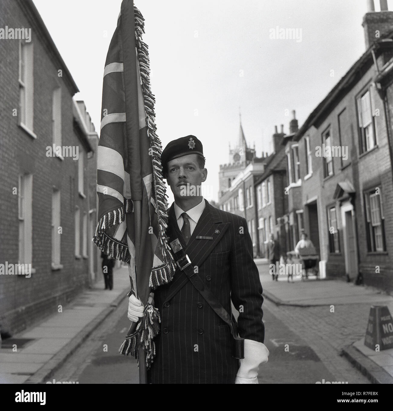 1964, Parade, ein Mann in einem Stift - gestreifte Anzug und das Tragen eines militärischen Stil Barett mit Abzeichen, und tragen weiße Handschuhe, stehend in einer Seitenstraße mit einer Stange mit einer Flagge, Aylesbury, Bucks, England, UK er Teil in einer Parade die Schlacht um England zu feiern, Prozessionen statt in englischer Marktflecken in dieser Ära der Opfer des britischen Militärs und der Öffentlichkeit während des Zweiten Weltkriegs gemacht haben. Stockfoto 1964, Parade, ein Mann in einem Stift - gestreifte Anzug und das Tragen eines militärischen Stil Barett mit Abzeichen, und tragen weiße Handschuhe, stehend in einer Seitenstraße mit einer Stange mit einer Flagge, Aylesbury, Bucks, England, UK er Teil in einer Parade die Schlacht um England zu feiern, Prozessionen statt in englischer Marktflecken in dieser Ära der Opfer des britischen Militärs und der Öffentlichkeit während des Zweiten Weltkriegs gemacht haben. Stockfoto