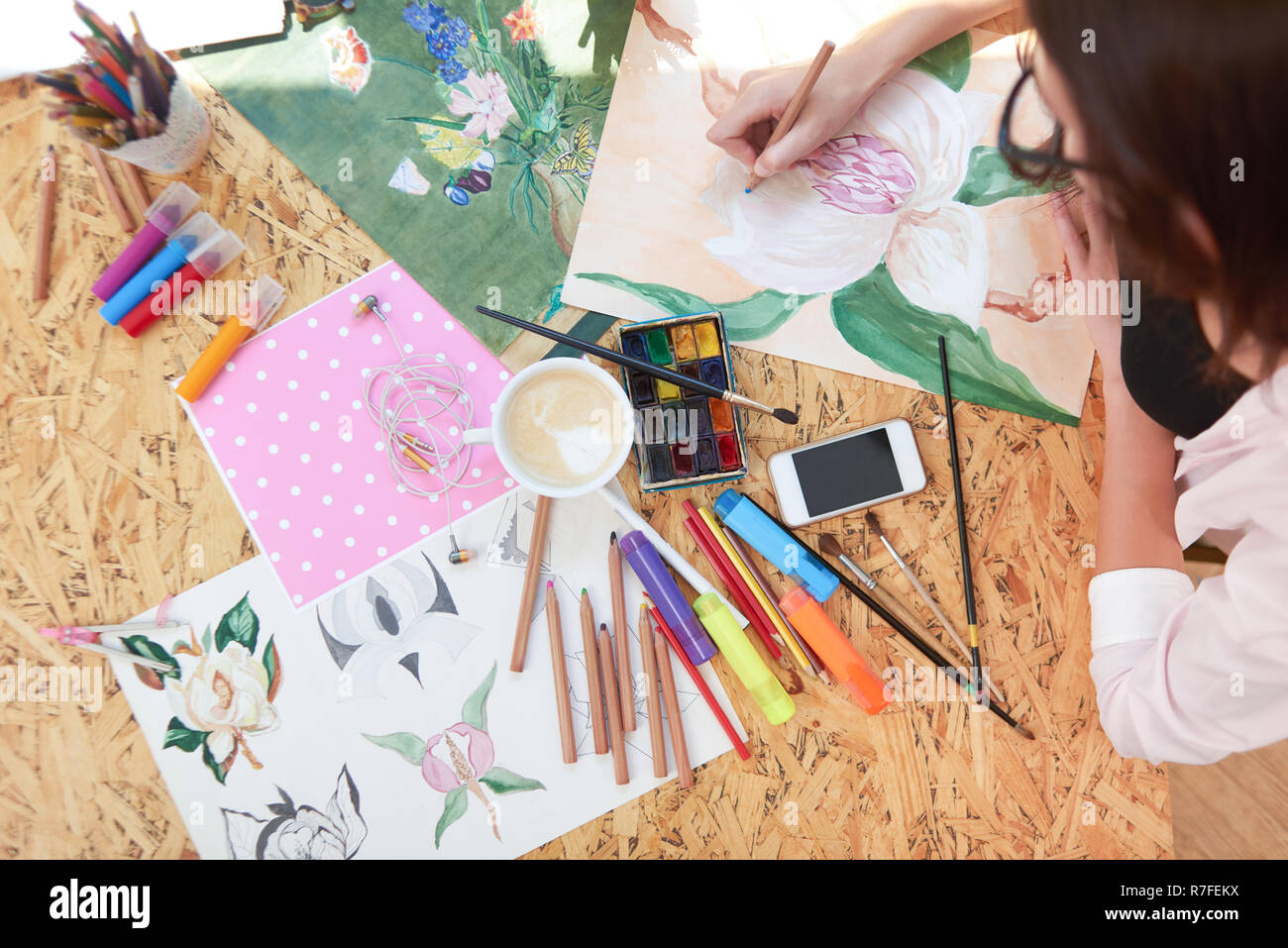 Blick von oben der Maler Zeichnung schöne Blume mit Bleistift in der Hand. Verschiedene Objekte als Container mit Bleistiften, Marker, Pinsel, Papier, Gadget und Tasse Kaffee auf Tisch. Stockfoto