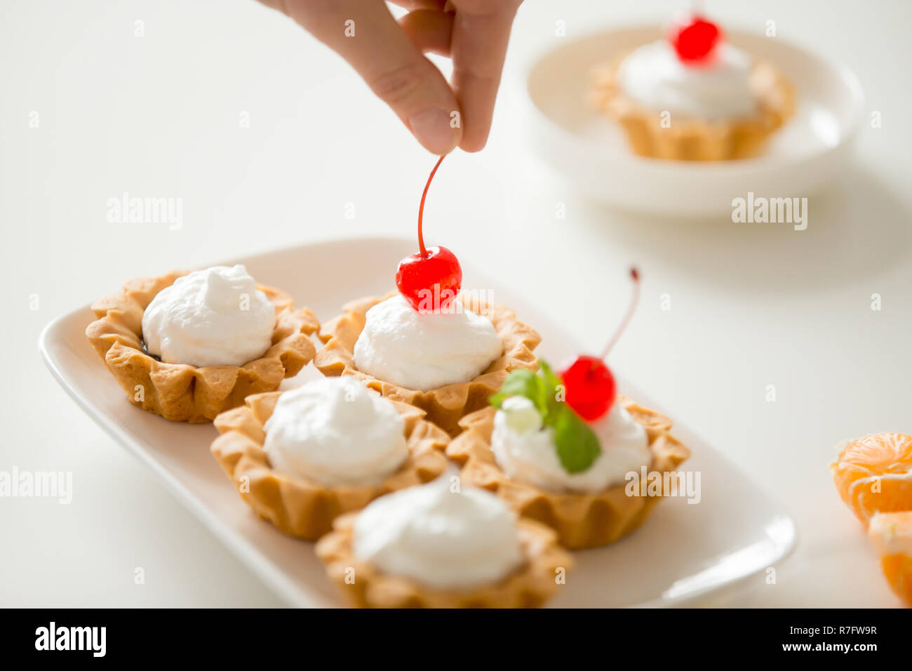 Nahaufnahme der Hand verzieren Bunte mini Obst und Sahne Torten mit roter Kirsche, in einer Reihe auf einem weißen Tisch Set Stockfoto