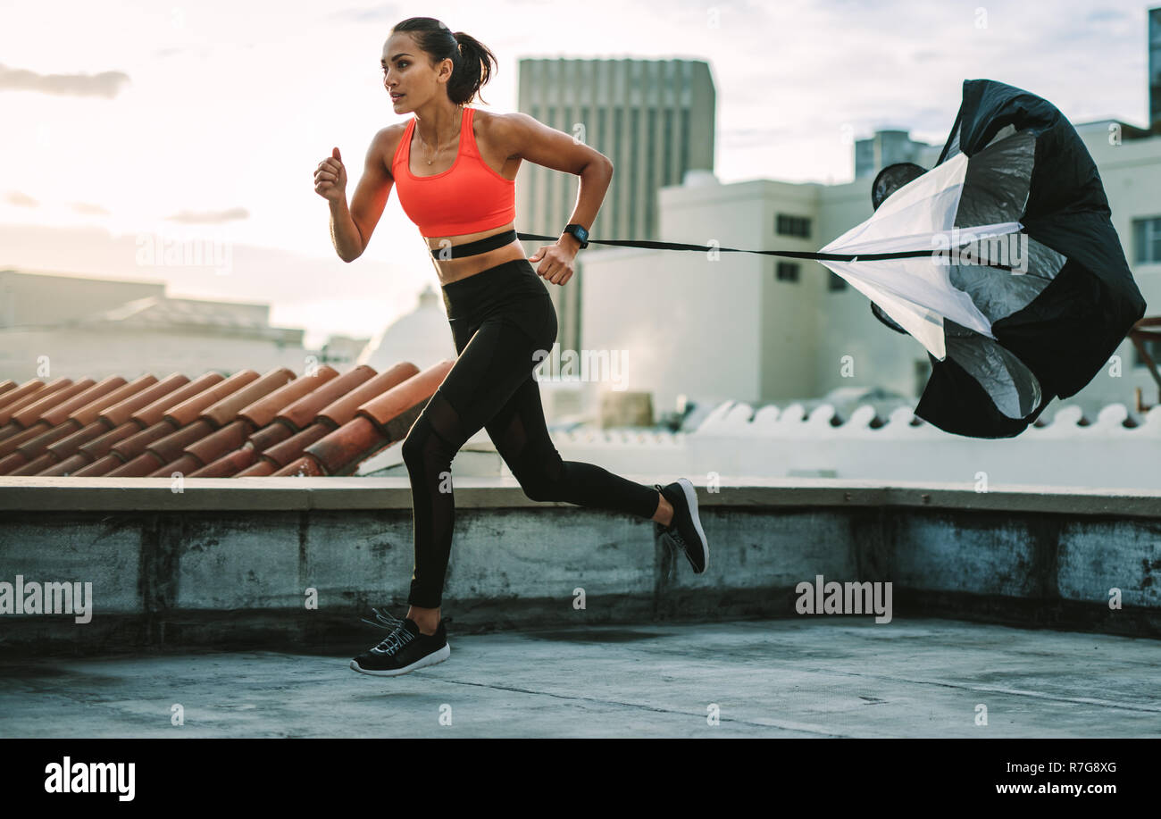Weibliche Athleten zu ziehen, die auf der Dachterrasse. Seitenansicht eines Fitness Frau läuft auf der Terrasse eines Gebäudes mit einem Widerstand Fallschirm zu ihr wa gebunden Stockfoto