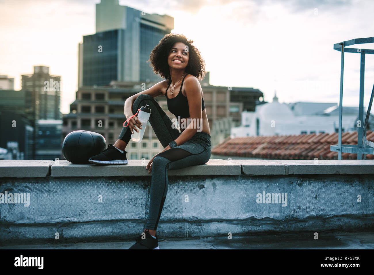 Lächelnd fitness Frau sitzt auf der Terrasse des Gebäudes Trinkwasser. Frau in fitness Verschleiß sitzen auf der Dachterrasse Zaun mit einem medizinball in der morn Stockfoto