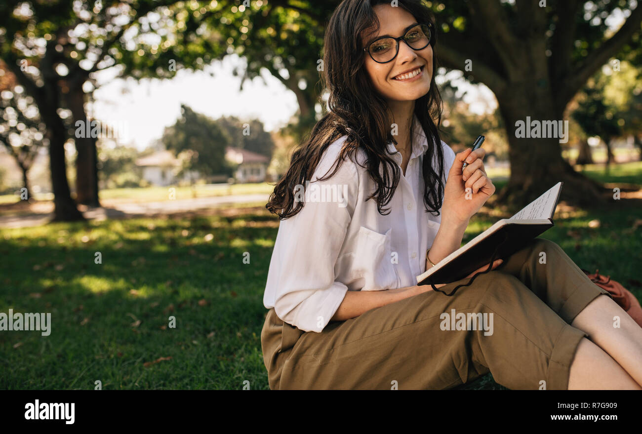 Schöne junge Frau mit Brille im Park schreiben in einem Buch sitzen. Kaukasische weibliches Schreiben in einem Buch im Park. Stockfoto
