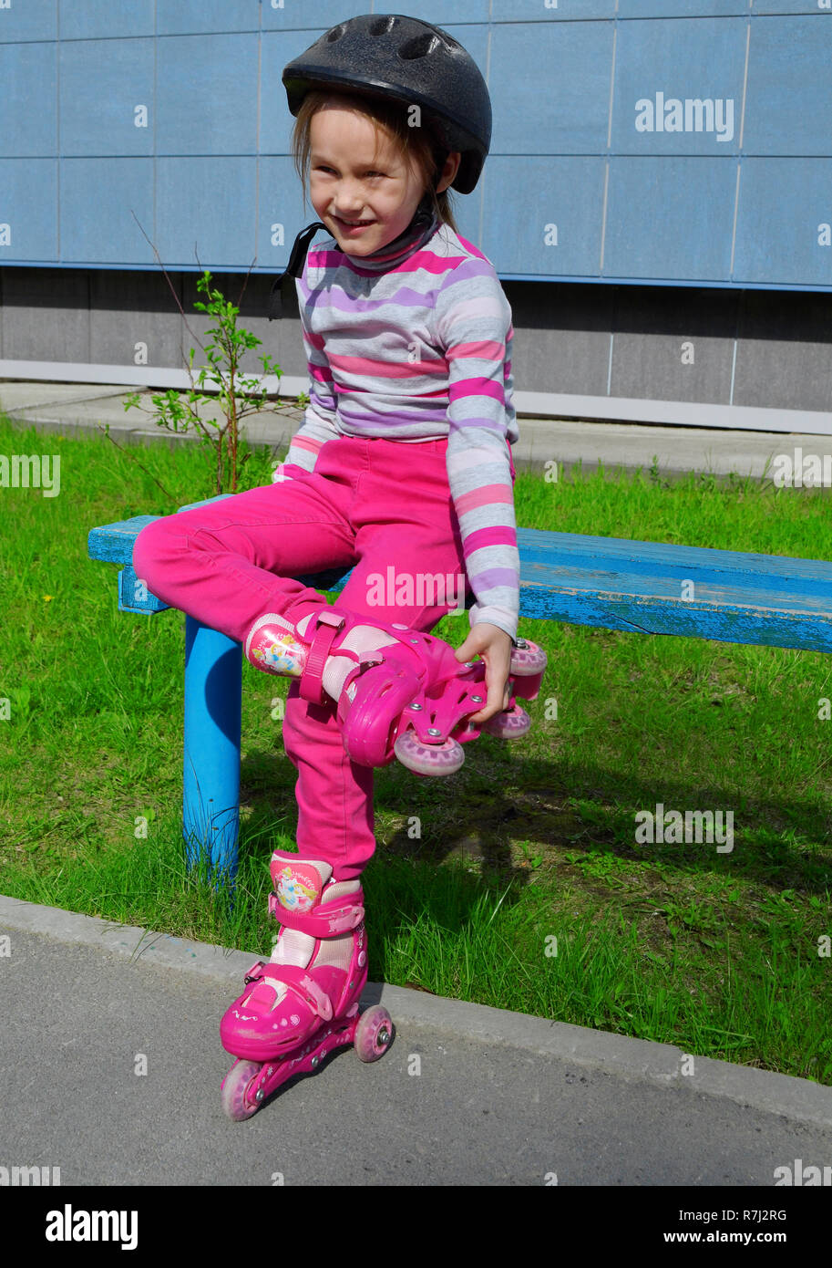 Ein Mädchen in rosa Jeans und einem gestreiften Pullover in einem Roller  Helm auf dem Kopf und der Rollen auf ihren Beinen sitzt auf einer Bank an  einem sonnigen Sommertag. Phot Stockfotografie -