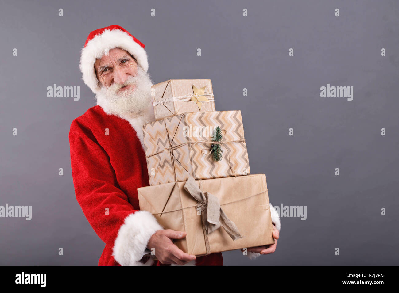 Santa Claus mit einem weißen Bart Tragen einer Brille und Santa Outfit trägt eine Menge Geschenke beim Stehen auf dem grauen Hintergrund, Silvester, Weihnachten, Feiertage, Souvenirs, Geschenke, Shopping, Rabatte, Geschäfte Stockfoto