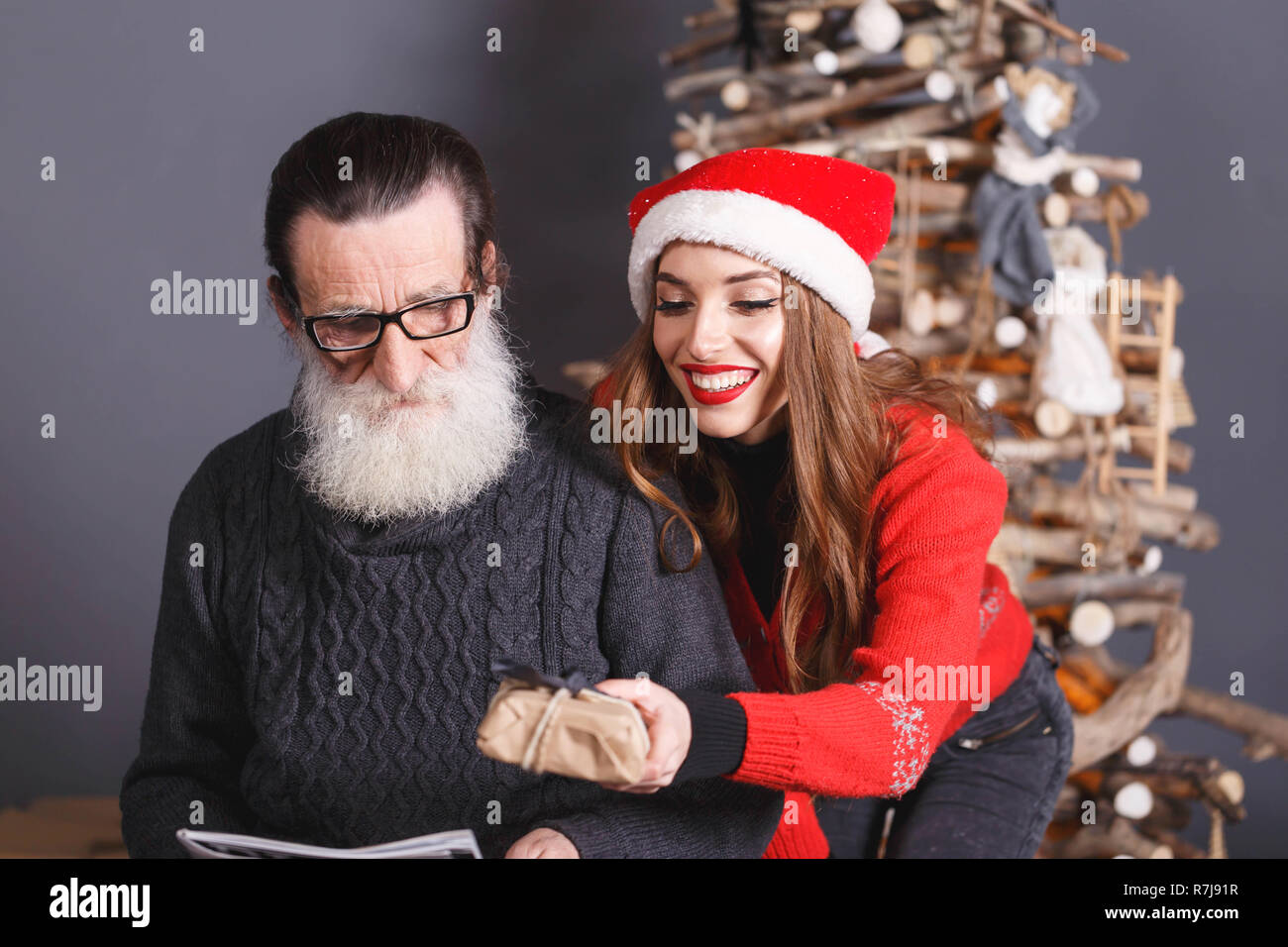 Attraktive langhaarigen Tochter im roten Pullover tragen Santa Hut gibt ein Geschenk zu Ihren bärtigen älteren Vater tragen graue Strickjacke, Silvester, Weihnachten, Feiertage, Souvenirs, Geschenke, Shopping, Rabatte, Geschäfte, Snow Maiden Santa Claus, Make-up, Frisur, Karneval. Stockfoto