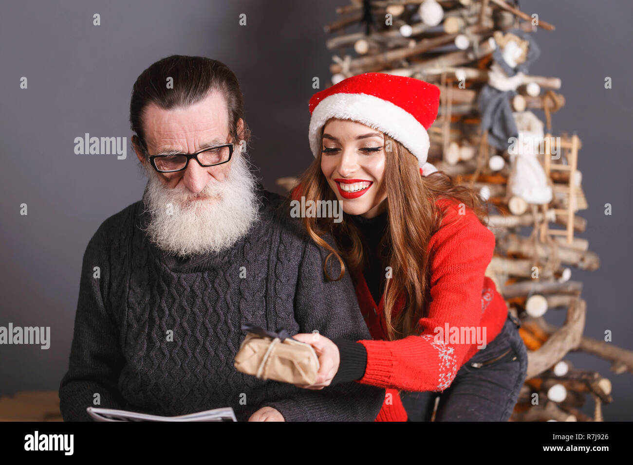 Die jungen langhaarigen Tochter im roten Pullover tragen Santa Hut lächelnd und gibt ein Weihnachtsgeschenk zu ihren bärtigen älteren Vati Brillen und graue Strickjacke, Silvester, Weihnachten, Feiertage, Souvenirs, Geschenke, Shopping, Rabatte, Geschäfte, Snow Maiden Santa Claus, Make-up, Frisur, Karneval. Stockfoto