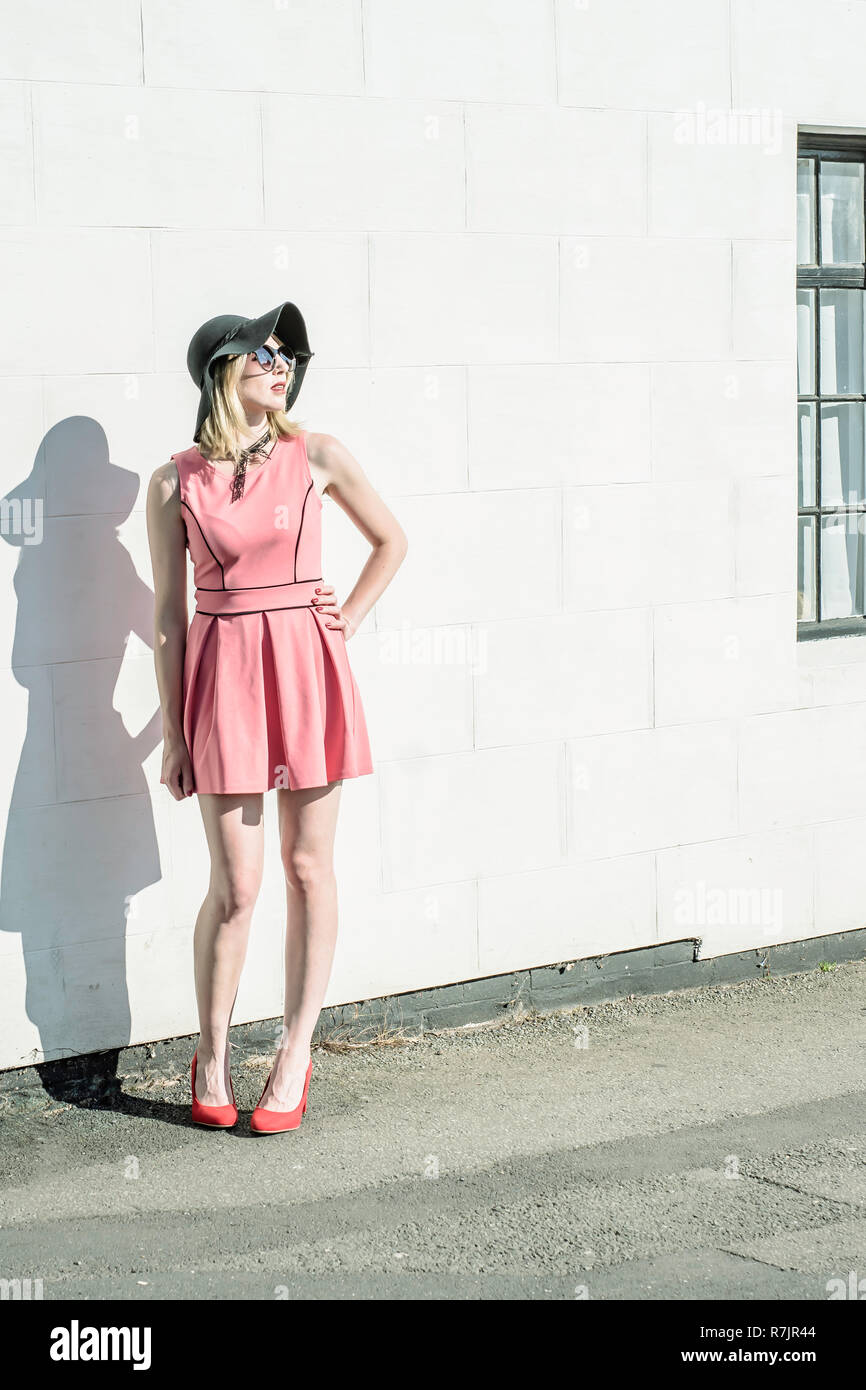 Authentische urban fashion. Junge, erwachsene Frau mit Sonnenbrille,  schwarzer Hut, rosa Kleid und rote Schuhe auf der Straße der britischen  Stadt im Sommer posieren. Weiß wa Stockfotografie - Alamy