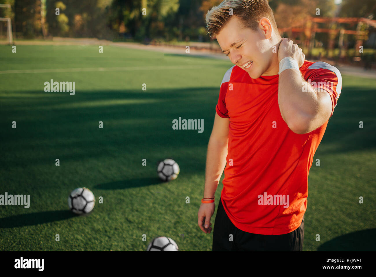 Junge Spieler fühlt Schmerz am Hals. Er halten es Hand und schrumpft. Kerl stehen auf Rasen. Die Kugeln sind hinter sich. Wetter ist gut und sonnig. Stockfoto