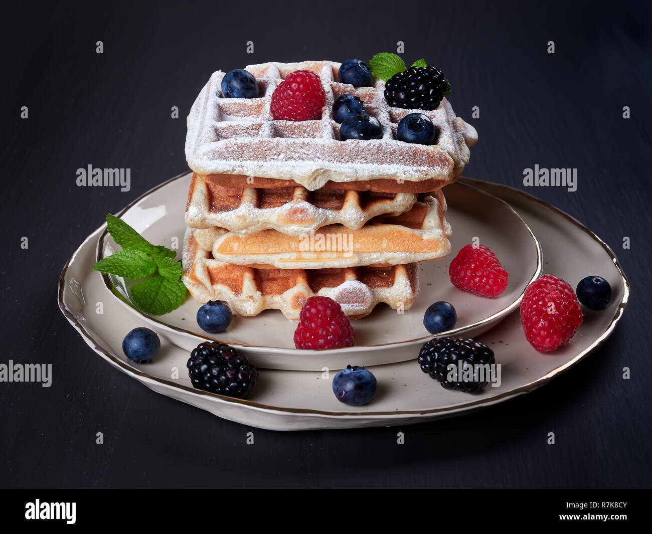 Ein Stapel mit vier home Belgische Waffeln mit Puderzucker bestäubt, mit frischen Beeren und Minze, auf eine beige Schüssel, auf dunklen braunen Hintergrund vorgenommen Stockfoto